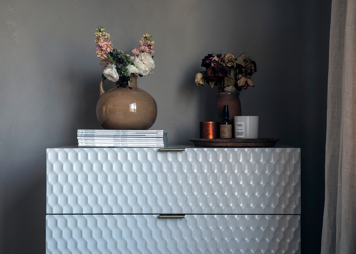 A white textured set of drawers styled with flowers, books, and candles atop.