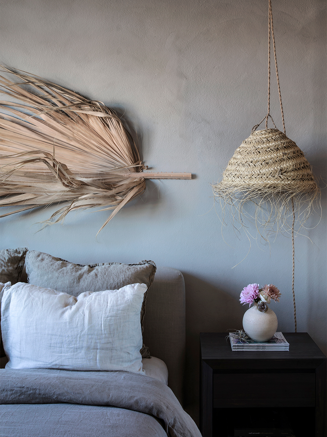 A bed with charcoal gray linen bedding against limewash wall and beside a woven pendant lamp.