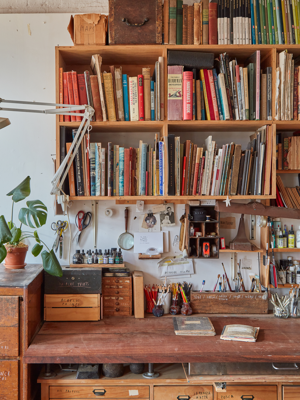 Antique wooden desk cluttered with pencils, paintbrushes, and art materials, below several rows of book shelves.