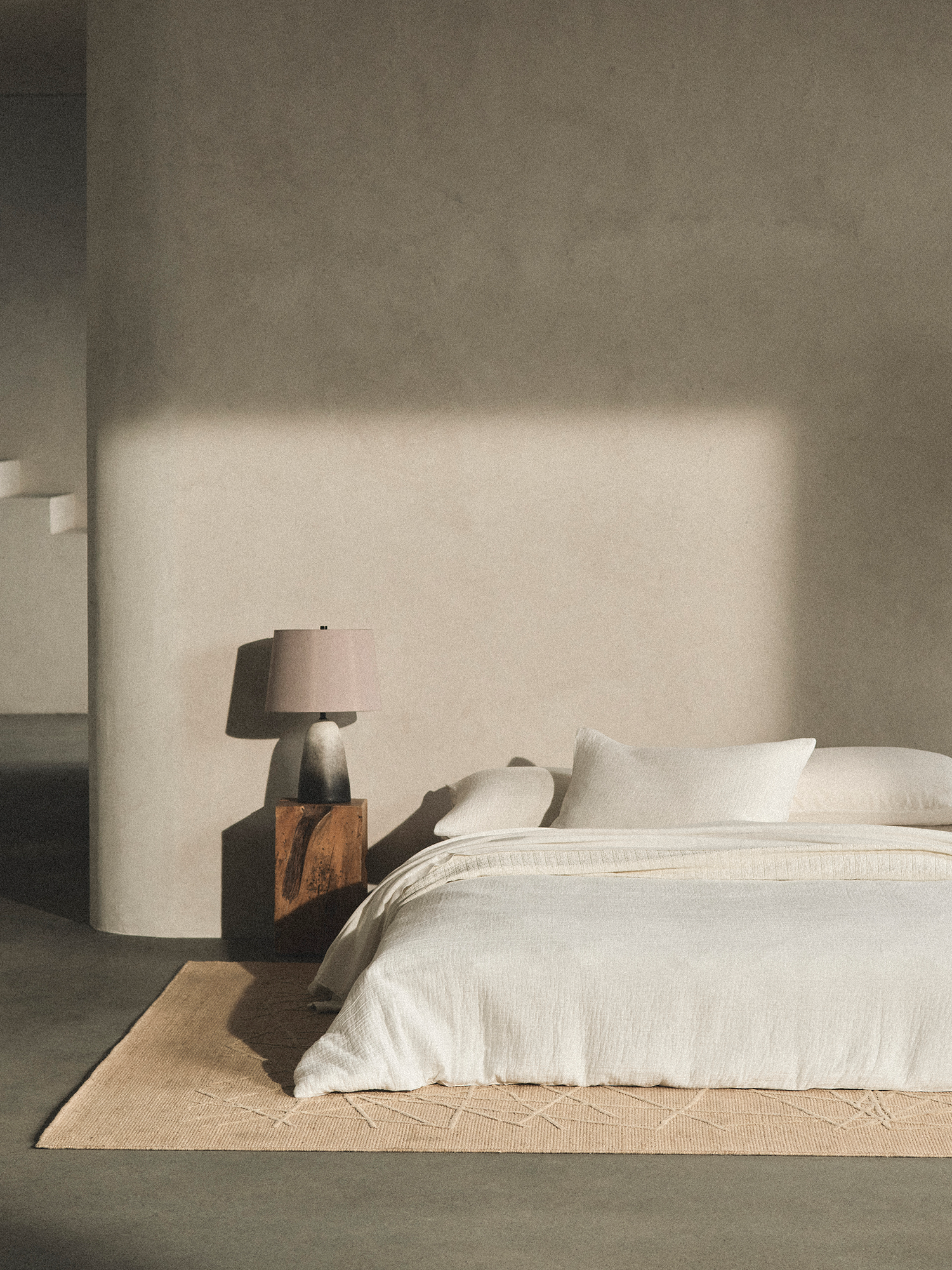 Sunlit bedroom with bed and wood bedside table against cream colored wall.